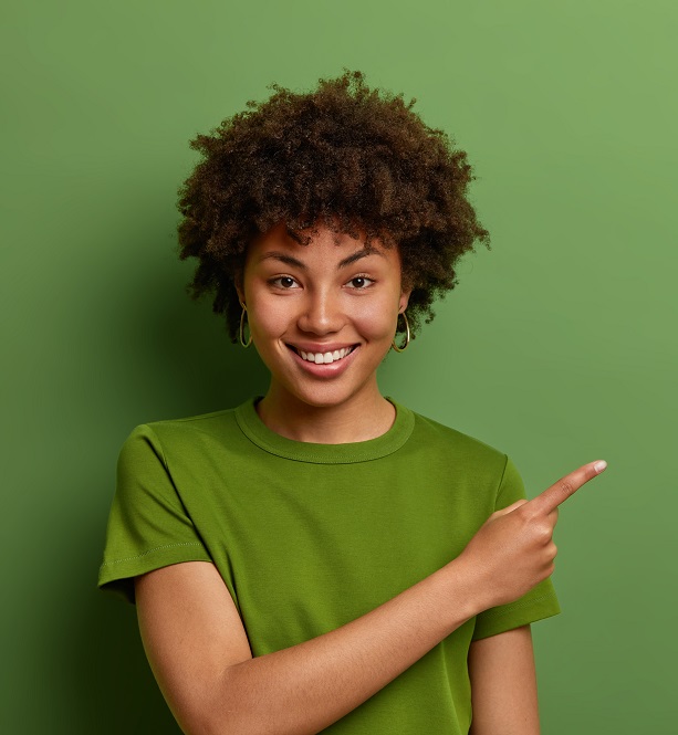 Friendly looking glad female shop assistant happy to help customers, shows way and demonstrates discounts in shop, points fore finger aside on empty space over green background. Advertising concept